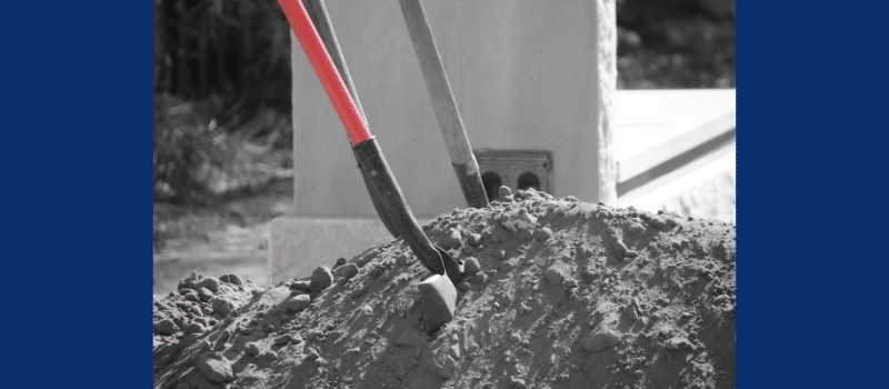 Two shovels stuck in a mound of dirt at a burial site, with a gravestone in the background.